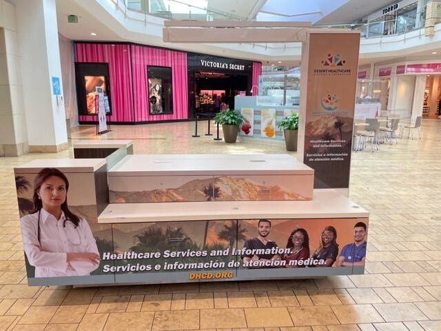 View of a kiosk in an interior corridor at Palm Desert Mall. The kiosk is adorned with images of diverse healthcare workers wearing reassuring expressions. Photo by Will Dean