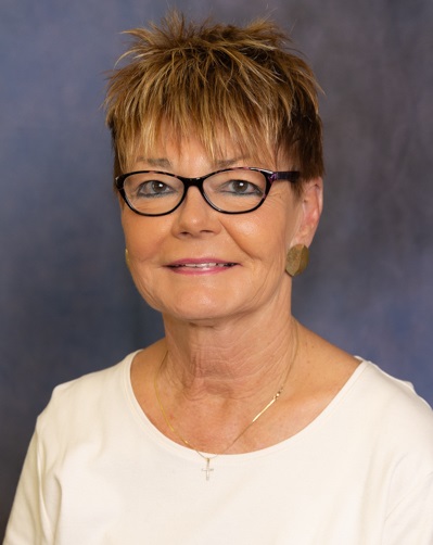 A headshot of Donna Craig facing the camera. She wear dark-framed eyeglasses, a white scooped-neck T-shirt. The background is a marbled purple and blue.