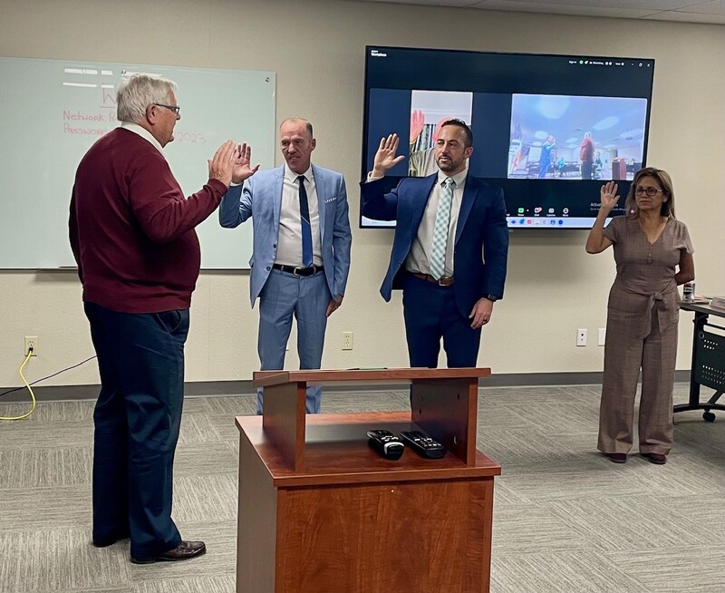 Attorney Jeff Scott stands with a raised hand facing three Board Directors who also have raised their right hands, in a conference room.