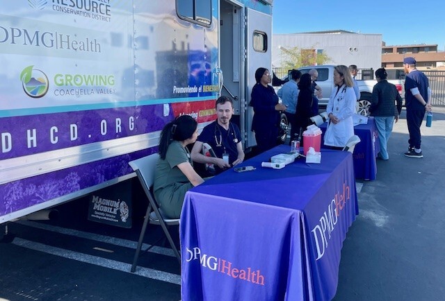 View of the parked mobile clinic with healthcare providers in scrubs seated and standing at two tables in front of the vehicle.