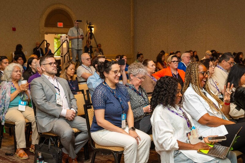 Side view of audience members seated in rows and looking toward an unseen stage, away from the camera.