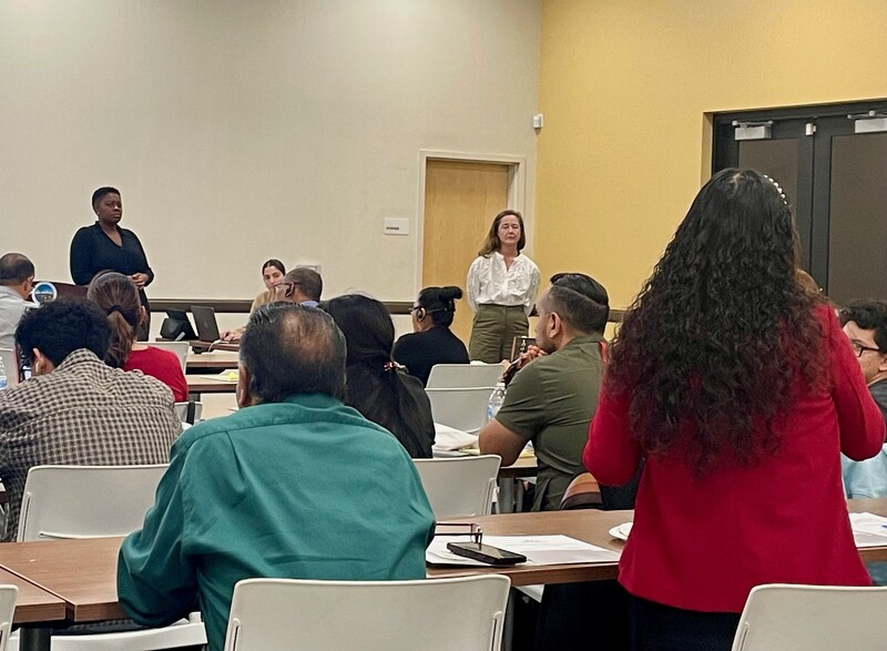 From the back of the room, the view shows the back of a woman who is standing and looking toward two women who are standing and facing her.