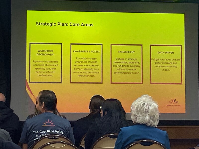 In the foreground, a seated audience from the back. They're looking at (in the background) a bright yellow-green screen that displays the four core areas of the strategic plan. Staff Photo